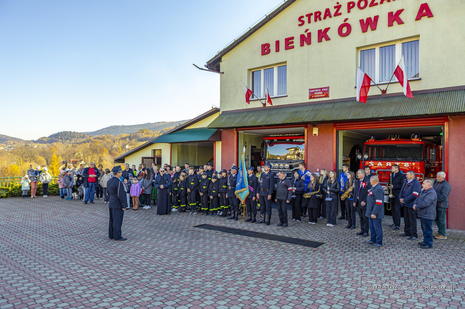 12:07. Remiza OSP Bieńkówka. Strażacy, poczet sztandarowy, orkiestra i mieszkańcy na placu.