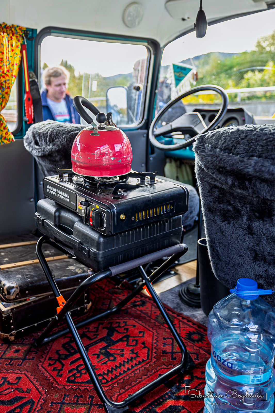 A Nysa van turned camper: curtains in the back, a camping stove with a kettle, a rug and a jug of water (2/2)