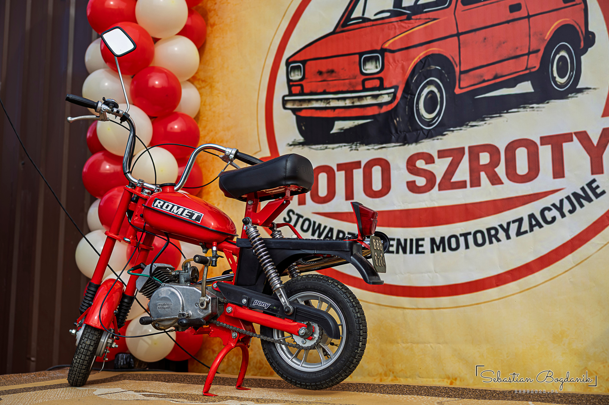 Czerwony motorower Romet przed banerem z rysunkiem malucha i napisem Moto Szroty.