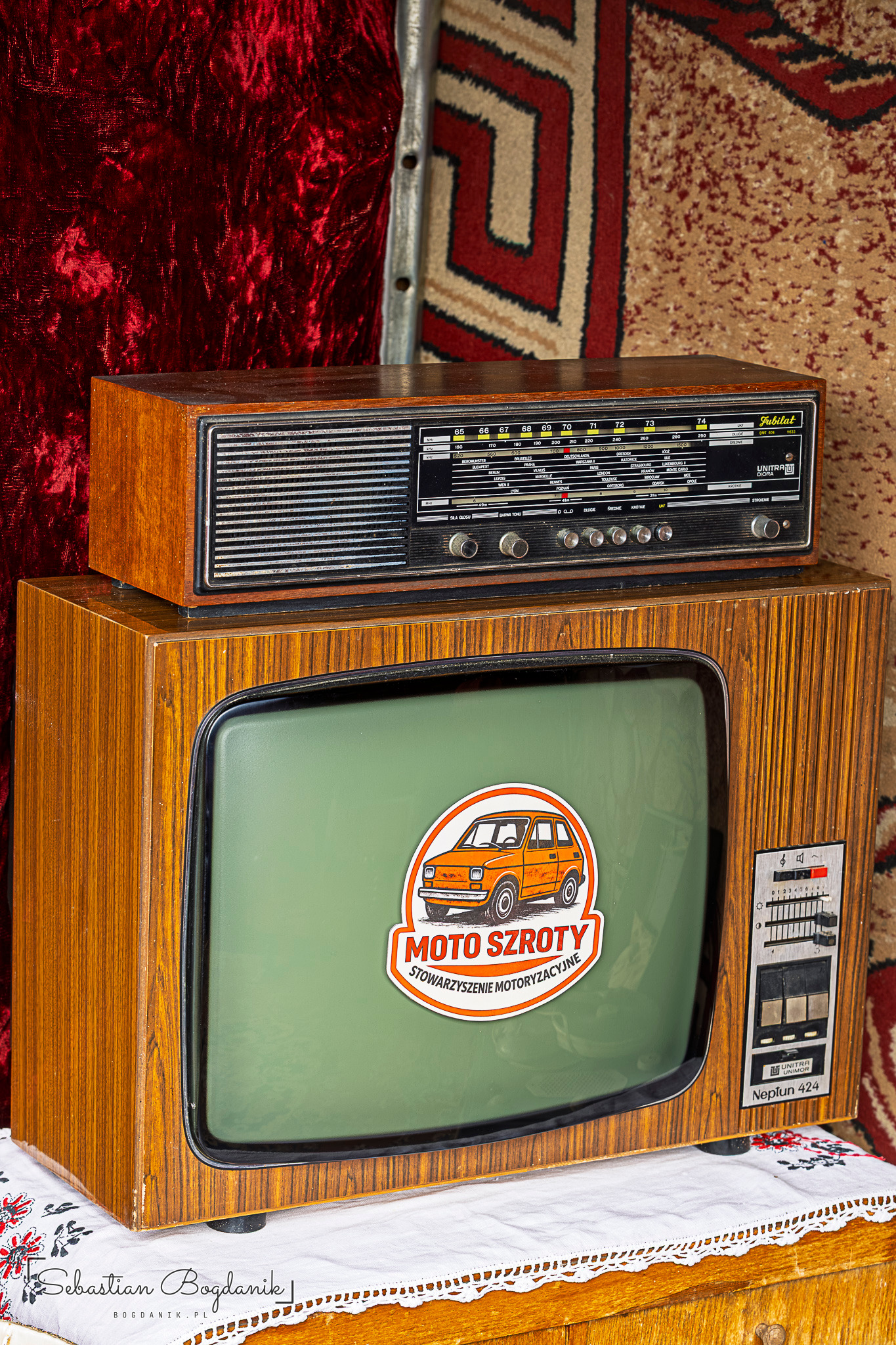 Drewniany telewizor z naklejką Moto Szroty na ekranie, na nim radio, w tle dywan we wzory.