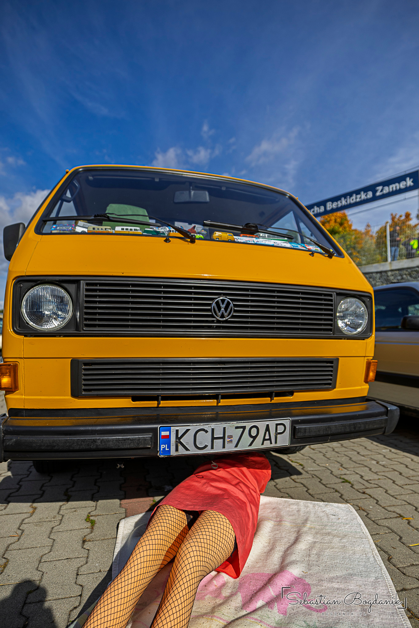 Żółty Volkswagen T3 od przodu, pod zderzakiem wystają nogi manekina w kabaretkach i czerwona sukienka.